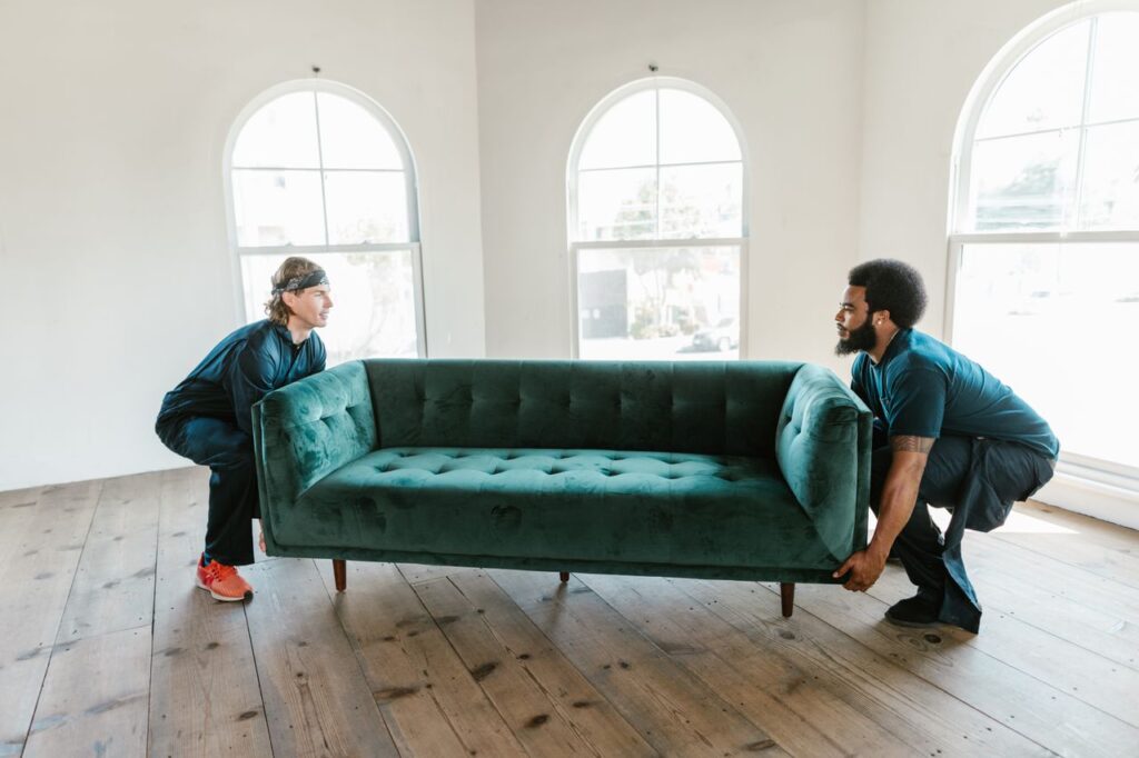 Two men carrying a green sofa in a sunlit room with wooden floors and arched windows.